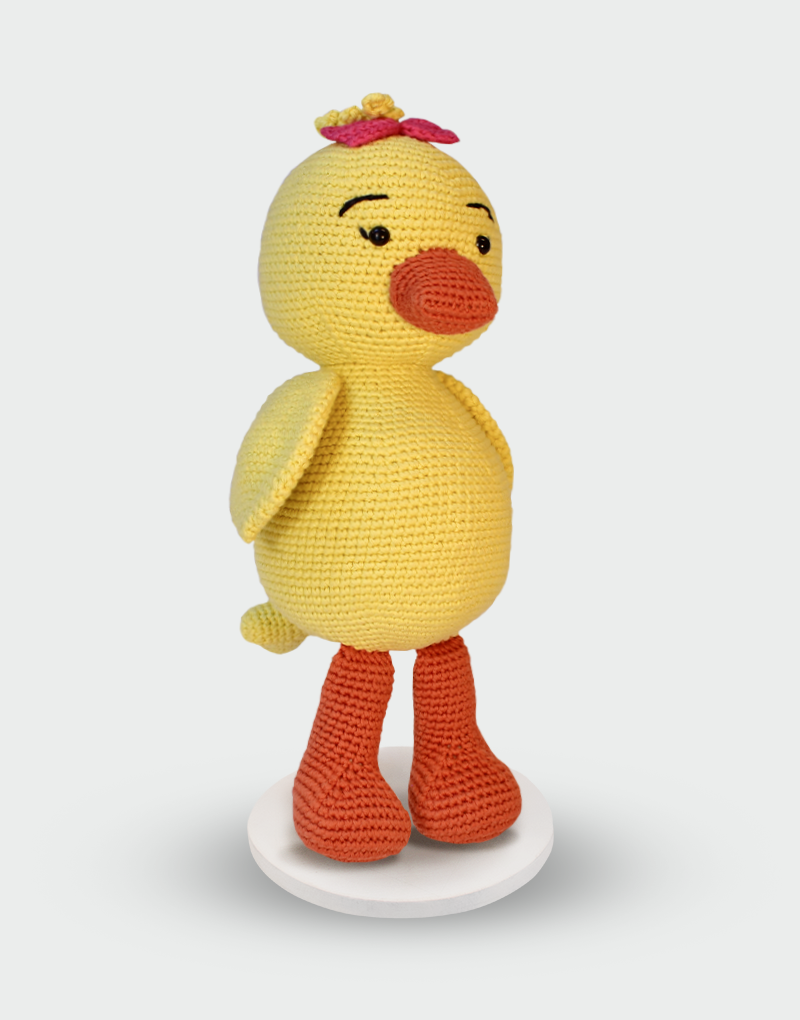 Muñeco amigurumi de pato amarillo tejido a mano en algodón orgánico con patas naranjas y lazo rosa en la cabeza sobre base blanca decorativa