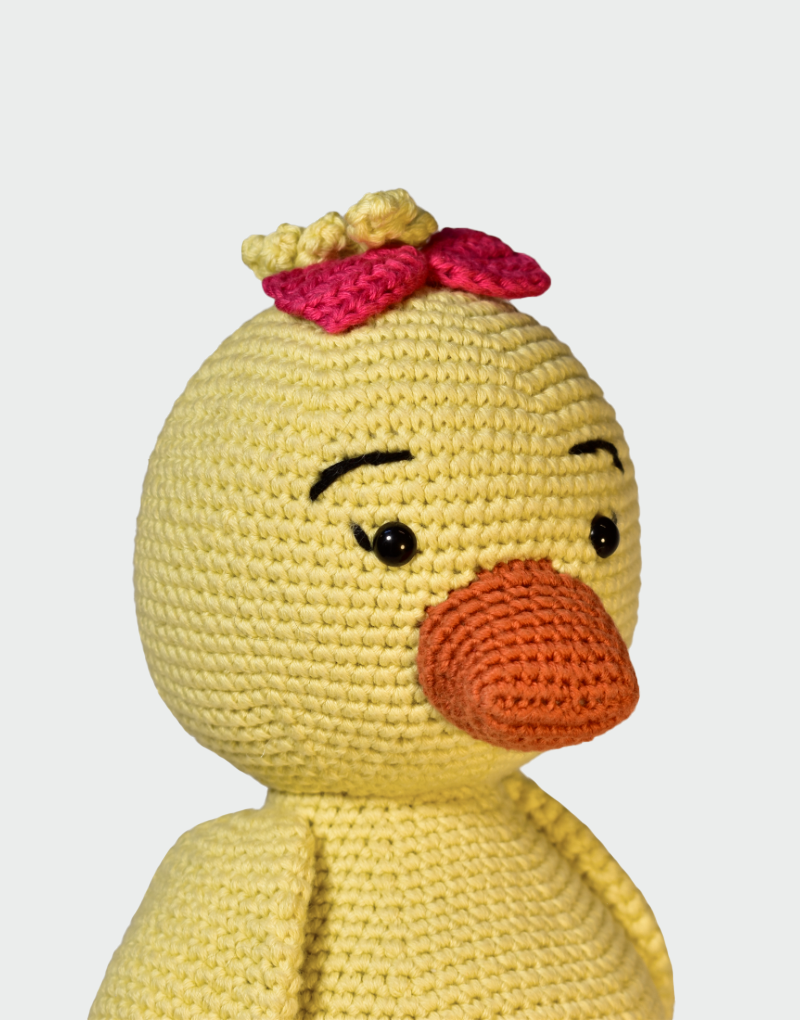 Primer plano de pato amigurumi tejido a mano en algodón orgánico amarillo con pico naranja ojos bordados y moño rosa decorativo en la cabeza