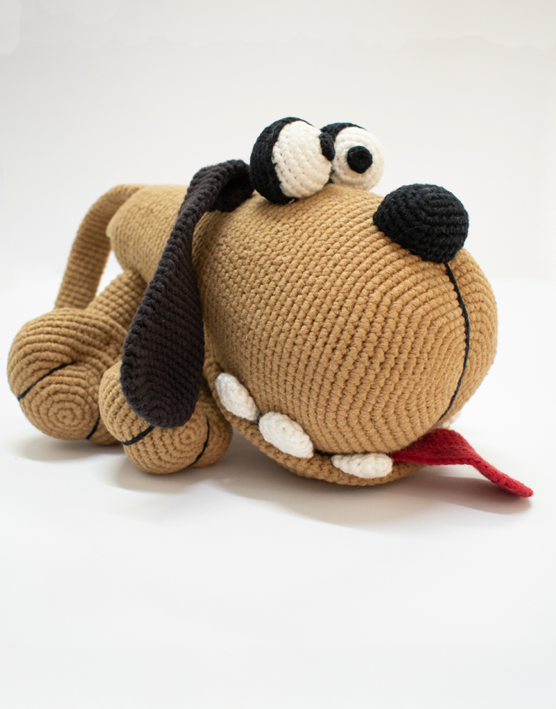 Juguete de peluche tejido a crochet con forma de perro marrón claro, orejas largas negras, ojos grandes y lengua roja, ideal para niños o decoración artesanal